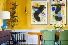 a quirky dining room with yellow walls, a bold gallery wall, a wooden table and green chairs, a retro chandelier and a potted plant