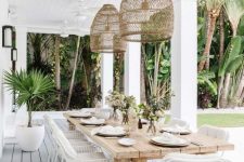 a relaxed tropical outdoor dining space with a stained wooden table, white chairs, pendant lamps and some greenery around