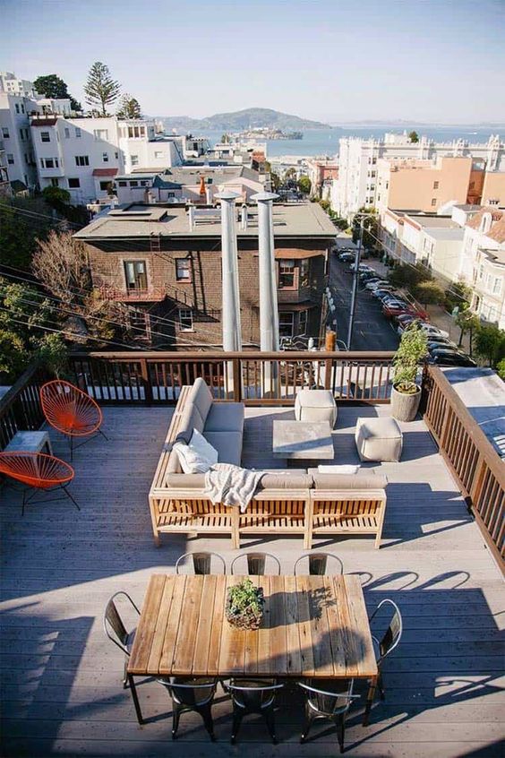 a rooftop terrace with a wooden deck, a sectional with poufs, bright chairs and a dining table plus metal chairs