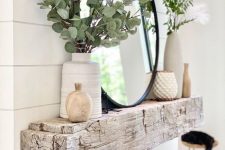 a rough wooden slab mantel with greenery arrangements in vases, elegant candleholders, a round mirror is a stylish idea