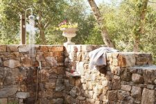 a rustic outdoor shower with stone walls, pebbles and tiles on the ground, potted greenery and blooms is a welcoming space