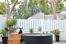 a simple and pretty backyard with a black stock tank pool, a wooden ladder and a deck, potted plants and fun floats is welcoming