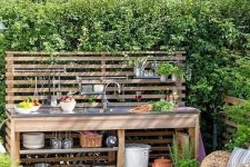 a small and cool outdoor kitchen with a planked screen used for attaching shelves, open shelves and a sink plus potted herbs
