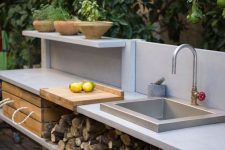 a small contemporary outdoor kitchen with an open shelving unit that stores firewod and a crate, with a built-in sink and potted herbs
