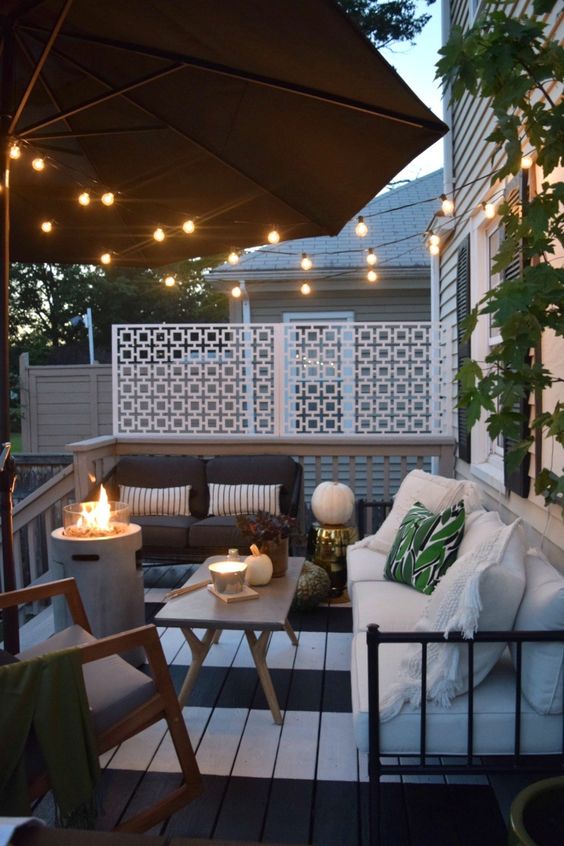a small deck with an umbrella and string lights, modern white and black furniture, a coffee table and a fire pit plus greenery