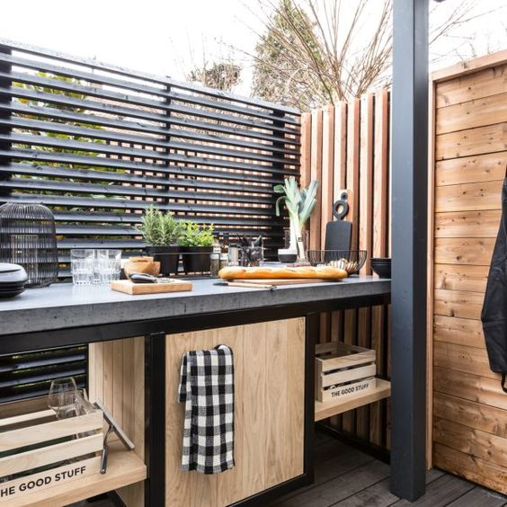 a small industrial and rustic outdoor kitchen with an open and closed storage unit, a concrete countertop, a planked wall and potted herbs