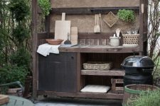 a small rustic outdoor kitchen of a single storage unit, with a roof, some shelves, a closed cabinet, potted greenery and cutting boards