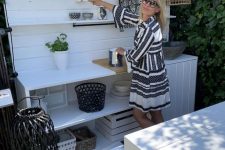 a small white outdoor kitchen with a cabinet and open shelves, railings, baskets and a pendant lamp plus a matching white planked dining set