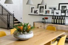 a stylish contemporary dining room with a large table, yellow chairs, grey pendant lamps, a dresser and a ledge gallery wall