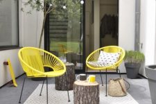 a stylish modern summer terrace with a printed rug, yellow chairs, tree stumps, potted greenery and a light string over the space
