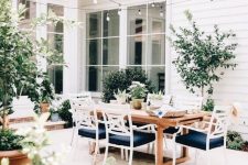 a stylish outdoor dining space with a sained table and white and navy chairs, potted plants and trees is a very cool space