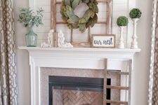a summer farmhouse mantel with greenery balls and a leaf wreath, a vintage window frame, some pillows and blankets