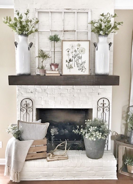 a summer mantel with greenery and flower arrangements, potted plants, a black and white artwork, a stack of books and a crate with pillows