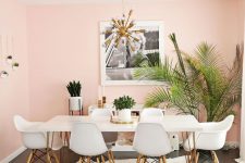 a tropical glam dining room with light pink walls, a hairpin leg dining table, white chairs, a sunburst chandelier and some tropical plants