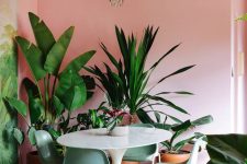 a tropical glam dining room with pink walls, a round table, green chairs, a whimsical chandelier and lots of potted plants