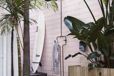 a tropical outdoor shower with a pink planed wall, a wooden deck, a surf board and a striped towel and tropical plants around