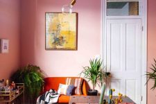 a vibrant dining room with pink walls, an orange loveseat, a dinind table with a colorful runner, some plants and a bar cart