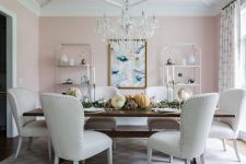 a welcoming refined dining room with blush walls, matching shelving units, a crystal chandelier, a wooden table and white upholstered chairs