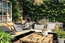 a welcoming summer terrace with a corner bench and printed pillows, a pallet coffee table, a pendant lamp and lights and potted greenery