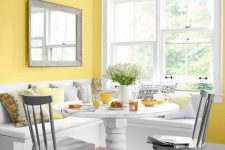 an airy country dining space with sunny yellow walls, a built-in bench and a white heavy table, grey chairs and bright printed pillows