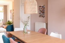 an eclectic dining room with dusty pink walls, a wooden dining table, pink, teal and white chairs and a woven pendant lam[