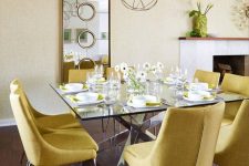 an elegant dining room with pale yellow walls, a glass table, yellow chairs, a mirror, a cluster of pendant lamps