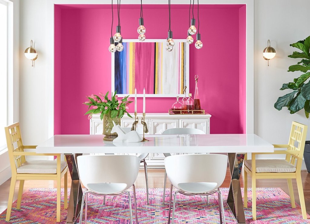 An eye catchy dining room with a hot pink niche with a bar, a long dining table and mismatching chairs plus bulbs over the space
