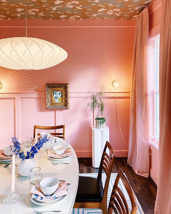 an eye-catchy dining room with pink paneled walls and a wallpaper ceiling, an oval table and stained chairs, a cool pendant lamp