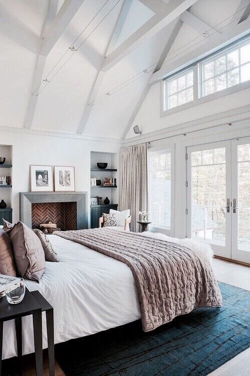 a beautiful and airy bedroom with a brick clad fireplace, a bed with white and dusty pink bedding, niche shelves, wooden beams