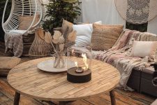 a boho terrace with cool wicker furniture and printed cushions and pillows, a white suspended chair on a stand, some string lights and a table fire pit