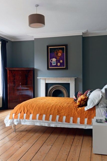 a bold and chic bedroom with grey walls, a fireplace, a dark stained dresser, a bed with bright bedding and a cool and bold artwork