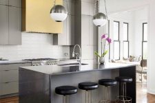 a bold contemporary kitchen with grey cabinets, a yellow hood, a grey ktichen island, chromatic fixtures, black stools with brass legs and pendant lamps with a silver finish