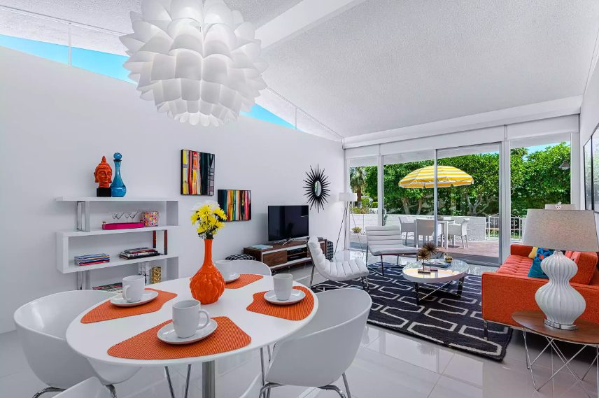 a bold living room with an orange sofa and a graphic rug, an oval table, white leather chairs, a TV unit and colorful artworks