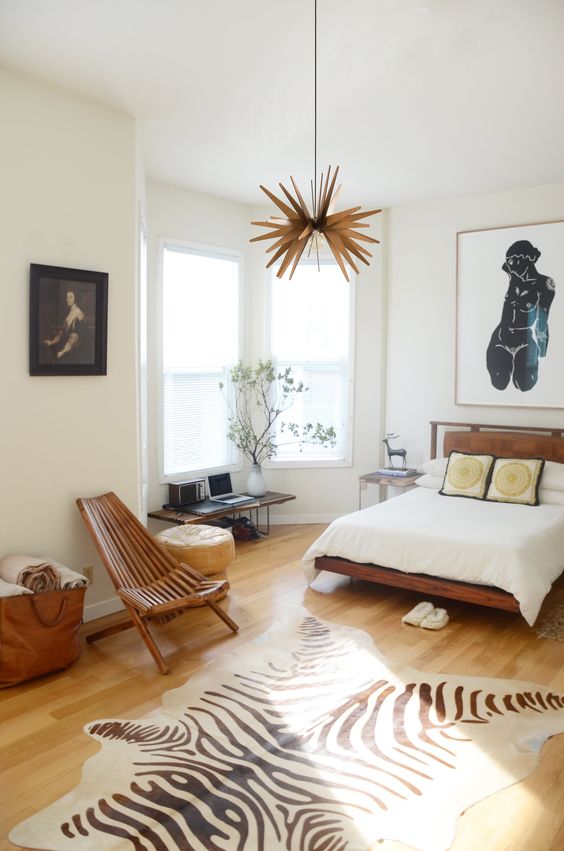 a catchy mid-century modern bedroom with a bay window, stained furniture, a faux animal skin rug, a pretty chandelier and cool art