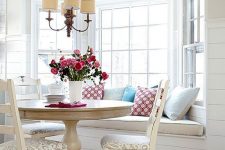 a charming cottage dining space with a bow window, a windowsill seating, a round table and neutral chairs plus a vintage chandelier