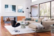 a chic mid-century modern living room with a tan sectional, a black chair, a glass table, a black floor lamp, a double-sided fireplace and cool art