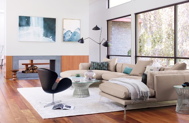 a chic mid century modern living room with a tan sectional, a black chair, a glass table, a black floor lamp, a double sided fireplace and cool art