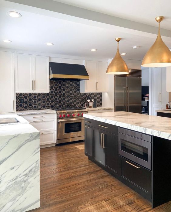 a chic modern kitchen with white shaker style cabinets, white stone countertops, a black hex tile backsplash, gold pendant lamps and chrome finish appliances