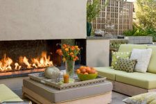 a chic modern outdoor space wiht a fireplace, neutral furniture wiht green and grey upholstery, a large ottoman and bold touches of orange
