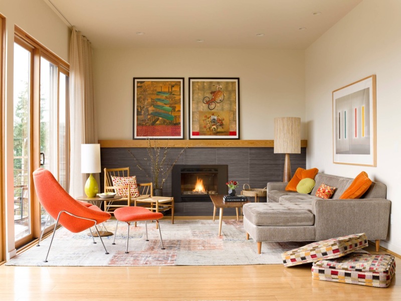 a colorful mid century modern living room with a built in fireplace, a grey sectional, a fiery red chair, a wooden chair, a low table and colroful artworks