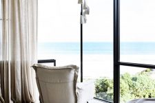 a contemporary bedroom with a neutral chair, a bed with neutral bedding and curtains plus a corner window to enjoy the sea views