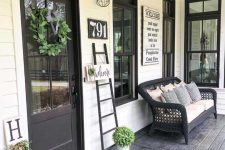 a contrasting rustic porch with a black door and deck, a black wicker sofa, a ladder, signs, potted greenery and sconces