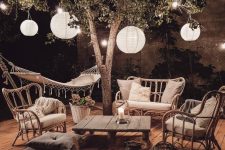 a cozy boho outdoor space with rattan furniture, a pallet table, woven candle lanterns, a small pouf and lots of paper lamps hanging around