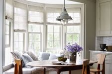 a cozy farmhouse nook with a bow window, a windowsill banquette seating, a wooden table and wooden chairs plus a retro lamp