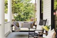 a cozy farmhouse porch with a black hanging daybed with lots of printed pillows, black rockers and lots of greenery
