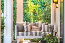 a cozy farmhouse porch with a hanging daybed with striped pillows, a wicker lounger, potted plants and blooms is a chic idea