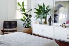 a cozy mid-century modern bedroom with a dark stained floor, a white storage unit, a bed with neutral bedding, statement plants and a black woven chair
