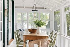 a cozy screened porch with a dining space – a wooden table, green and natural rattan chairs, greenery and a pendant lamp
