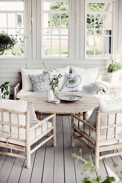 a dreamy Nordic veranda with bamboo furniture including a round table, neutral and printed bedding, potted greenery is wow