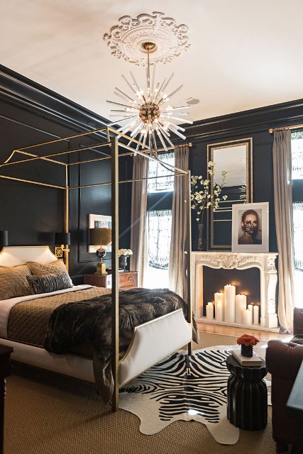 a fantastic glam bedroom with black walls, a gold canopy bed with pretty bedding, a sunburst chandelier, a fireplace wiht candles and layered rugs